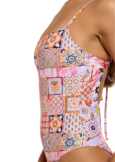 Side view of woman facing left wearing the colorful La Mer One Piece swimsuit with thin straps, featuring a vibrant patchwork of floral and geometric patterns, including fruits, flowers, tiles, and the word "la mer" on pink background, left armpit, chest to hips visible, on white backdrop.